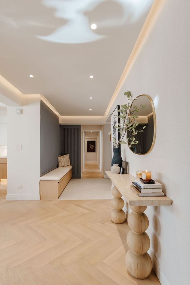 A refined residential hallway and entry area with pale herringbone timber flooring, warm white walls, recessed ceiling lighting, and concealed cove lighting. A built-in dark-panelled bench with drawers sits along the left wall, while a sculptural light timber console with spherical supports, an oval brass-framed mirror, books, candles, and a tall vase create a softly luxurious focal point on the right. The corridor extends toward a darker artwork at the rear, with a partial glimpse of an adjacent kitchen or living space at left.