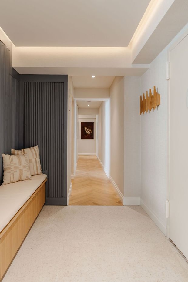 A refined interior hallway or entry area viewed toward a long corridor. The foreground has pale speckled stone or terrazzo-style flooring, white walls, recessed ceiling lighting, and concealed cove lighting. A built-in timber bench with cream upholstered seating and patterned cushions sits beside a dark charcoal vertical-ribbed feature wall or screen. The corridor beyond has warm pale timber herringbone flooring, white trim, and a small artwork at the far end. A sculptural timber wall object adds a warm accent on the right.