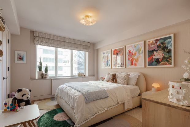 A warm, softly lit bedroom with a double bed centered against a pale upholstered headboard, layered white bedding, and neutral patterned cushions. A large window with a Roman blind overlooks nearby apartment buildings, while pale built-in cabinetry sits beneath it. Framed floral artwork forms a gallery wall above the bed, complemented by a light timber-toned bedside table, dresser, children's toys, a plush panda, and a decorative ceiling light. The room has a refined, calm, contemporary-classic feel.