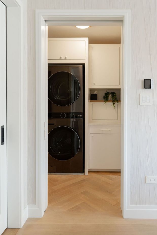 A compact laundry room viewed through an open doorway, featuring stacked black front-loading washer and dryer, pale cream built-in cabinetry, a small open shelf with a trailing plant, and warm herringbone timber flooring. The surrounding hallway has white textured wall panelling, detailed white door trim, a wall control panel, and light switches. The space is clean, bright, and refined with a warm neutral palette.