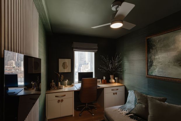 A compact, dark-toned home office with a built-in desk spanning the window wall, white drawer cabinetry, a brown office chair, and a large sofa in the foreground. Vertical timber wall slats, deep green textured walls and ceiling, warm timber flooring, framed artwork, decorative objects, and a ceiling fan with integrated light create a moody, refined workspace. City buildings are visible through the windows.