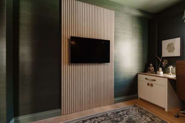 A moody, refined media or living room featuring a wall-mounted television set into a vertical timber-slat feature wall. Dark textured green walls surround a cream built-in sideboard with decorative hardware, while a patterned rug, artwork, sculptural decor and warm timber flooring add depth. The space has a low-contrast, dimly lit atmosphere with layered natural and dark neutral tones.