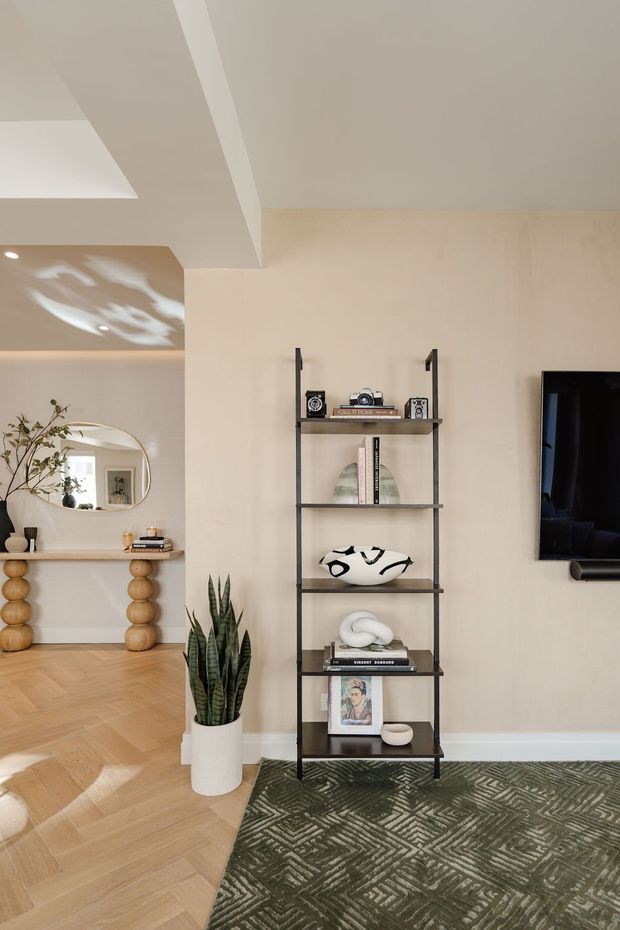 A refined contemporary living room with warm beige plaster walls, a white ceiling, and light herringbone timber flooring. A slim black metal open shelving unit displays books, ceramics, artwork, and decorative objects beside a wall-mounted television. In the adjoining area, a rounded mirror hangs above a narrow console with sculptural wooden sphere legs. A tall snake plant and patterned dark rug add contrast, while soft natural light creates a calm, curated atmosphere.