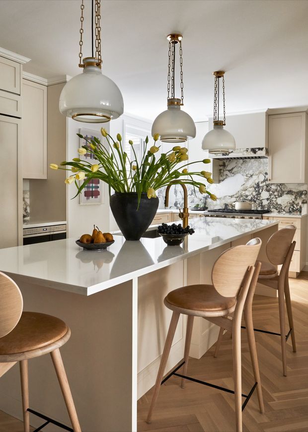Bright, refined kitchen interior with a large pale island, integrated cabinetry, marble-look veined stone splashback and benchtops, and three oversized opal-glass pendant lights suspended on decorative chains. Curved timber bar stools, a dark sculptural vase with yellow flowers, fruit bowls, brass-toned tapware, and herringbone timber flooring create a warm, elegant contemporary-classic atmosphere.