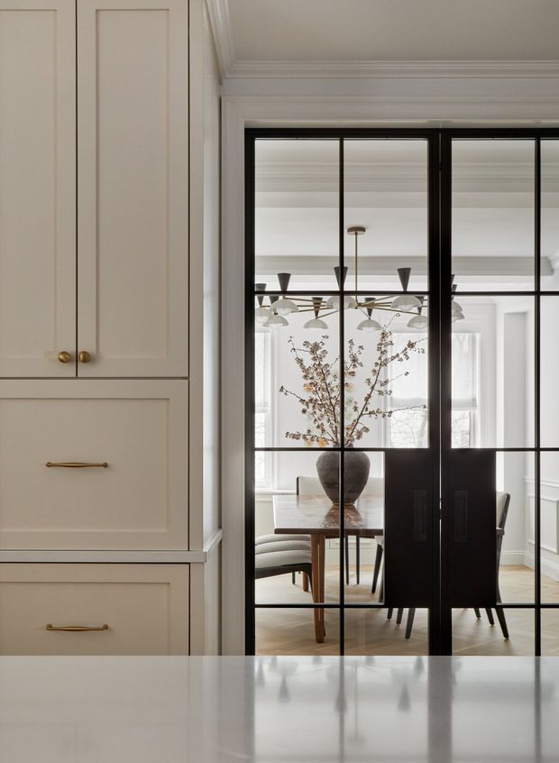 View from a kitchen toward a refined dining room through black-framed glazed steel-style doors. The foreground features tall cream built-in cabinetry with paneled fronts and brass hardware, while the adjoining bright dining room has a timber table, upholstered seating, a sculptural pendant light, and a large vase with branches. Ornate ceiling cornices and pale walls contrast with the dark metal-framed doors.