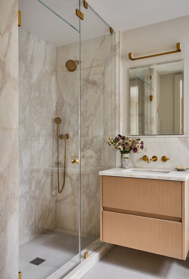 A refined contemporary bathroom featuring a frameless glass walk-in shower with large beige and cream marble-look wall tiles, small light mosaic floor tiles, and brushed brass shower and tapware. A floating light-wood vanity with a white stone benchtop sits beneath a large mirrored cabinet, complemented by a horizontal brass-accented wall light. The space has a warm, luxurious and carefully coordinated finish.