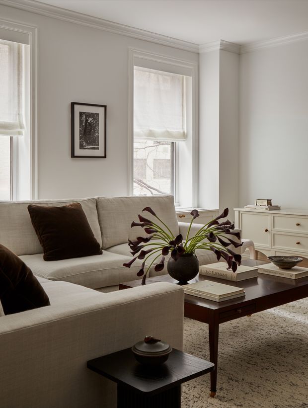 A refined, softly lit living room with two light neutral upholstered sofas arranged around a dark rectangular timber coffee table. The room features pale walls, tall sash-style windows with sheer Roman blinds, detailed white trim and cornices, a white built-in or freestanding dresser, framed artwork, a textured neutral rug, and restrained dark wood and burgundy accents. The styling is calm and contemporary with traditional architectural detailing.