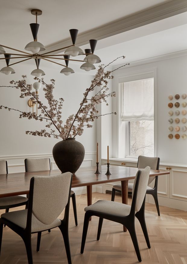 Elegant dining room with a long timber dining table surrounded by upholstered dark-framed chairs, a large sculptural vase of flowering branches, and a dramatic multi-arm pendant chandelier. The room features warm herringbone timber flooring, pale moulded wall panelling, decorative cornices, a tall window with a Roman blind, and understated neutral decor.
