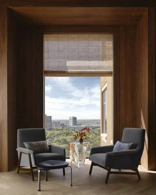 A refined seating nook framed by dark timber wall panelling and deep window reveals. Two charcoal upholstered lounge chairs face one another across a faceted glass side table with dried foliage, while a woven Roman blind filters daylight above the expansive city and tree-canopy outlook. The room has a warm, sophisticated and quietly luxurious atmosphere.
