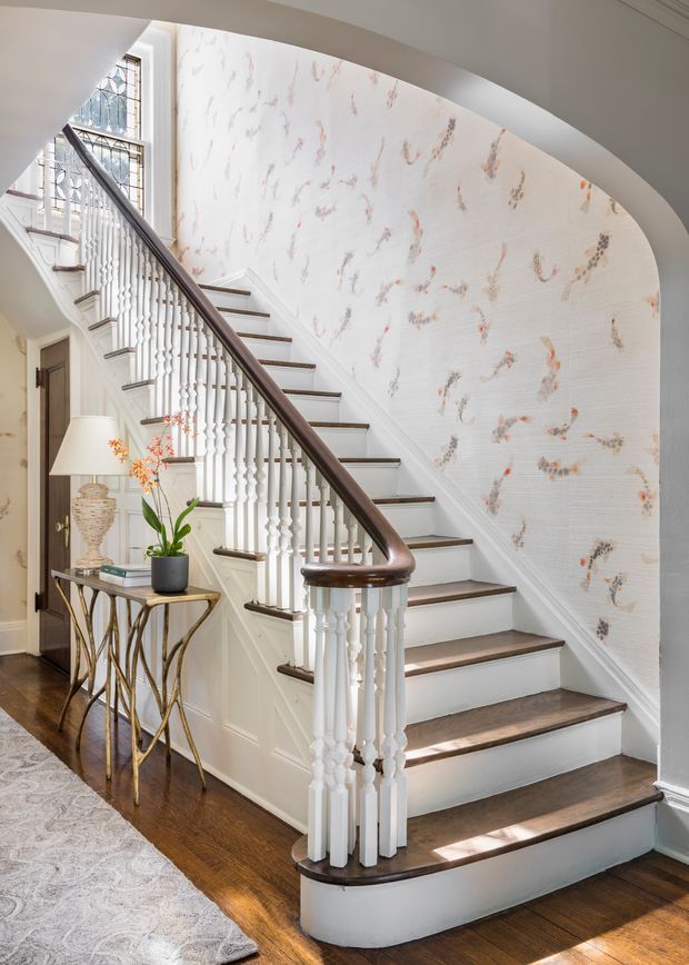 A bright, elegant interior stairway with a curved staircase, white turned balusters, dark timber handrail and stair treads, and white painted trim. The stair wall features delicate botanical-patterned wallpaper, while a slim console table, lamp, plant, and framed artwork sit beneath the stairs. Dark timber flooring and a pale patterned rug complete the refined traditional setting.