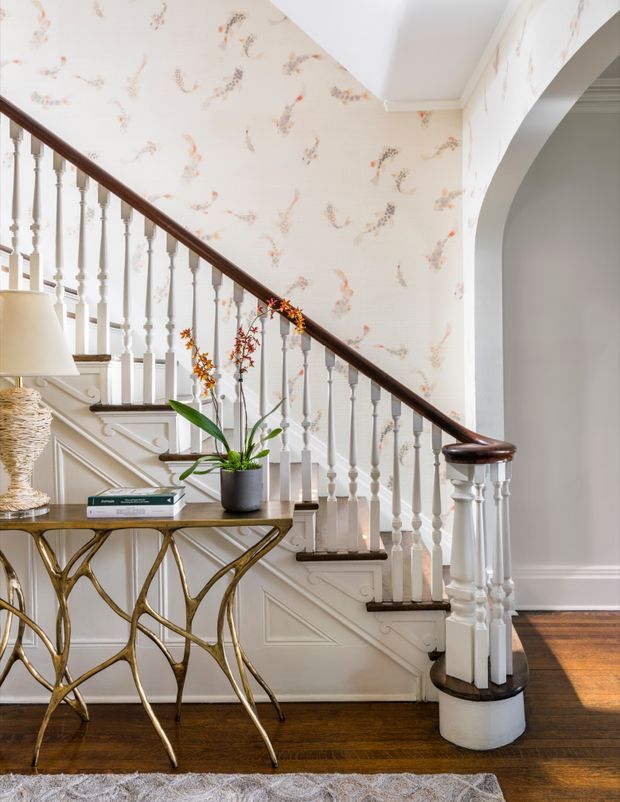 A refined residential stairway and landing with a traditional painted timber staircase, turned white balusters, dark wood handrail, panelled stair risers and skirting, and a large arched opening. Delicate floral wallpaper covers the walls. A small sculptural console table, lamp, books and potted plant sit beneath the stairs, with dark timber flooring and a partially visible rug.