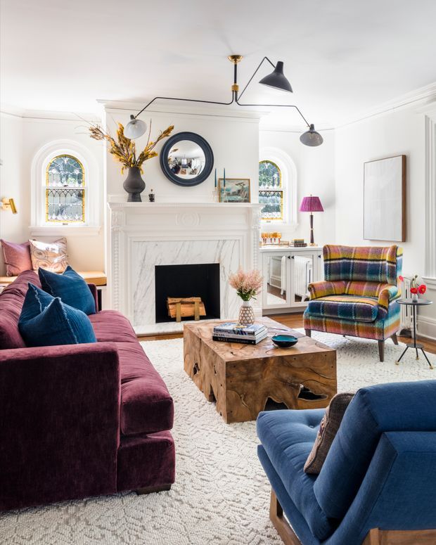 A bright, richly layered living room with a white painted fireplace mantel, marble-look surround, arched stained-glass windows, and ornate ceiling mouldings. The room features a deep burgundy velvet sectional, blue velvet armchairs, a multicoloured plaid lounge chair, a rustic live-edge timber coffee table, patterned pale carpet, framed artwork, and sculptural black pendant lighting. The overall look is eclectic, colourful, and refined within a traditional architectural shell.