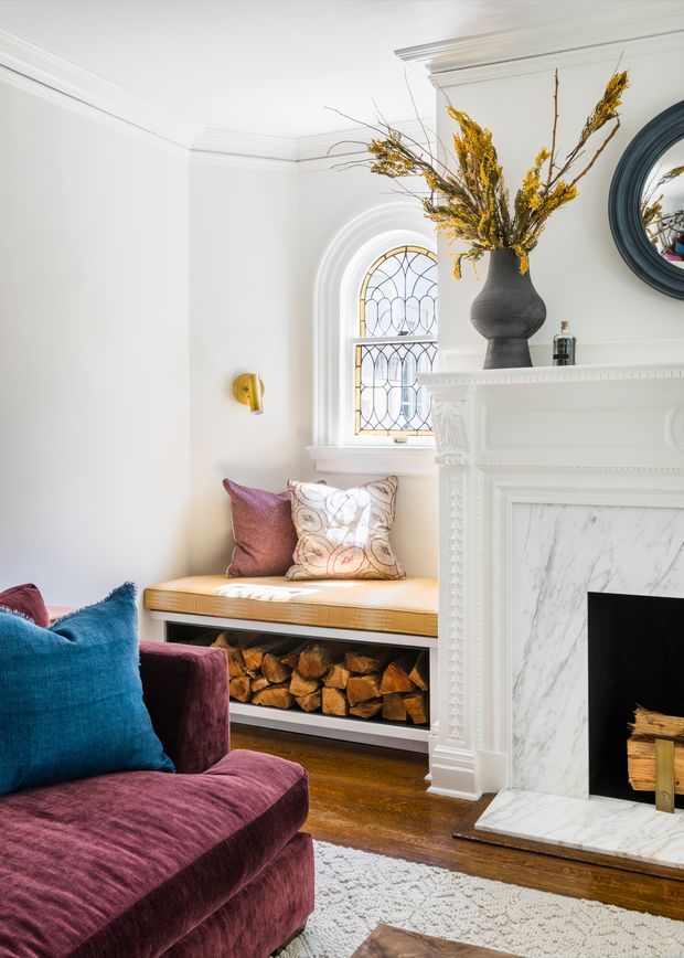 A refined living room corner with a white decorative fireplace mantel, marble surround and hearth, arched leaded-glass window, built-in window seat with patterned cushions, and integrated firewood storage. A deep plum velvet sofa with vivid blue cushions occupies the foreground, while a dark sculptural vase and dried floral arrangement decorate the mantel. Dark timber flooring and a pale textured rug add warmth and contrast.
