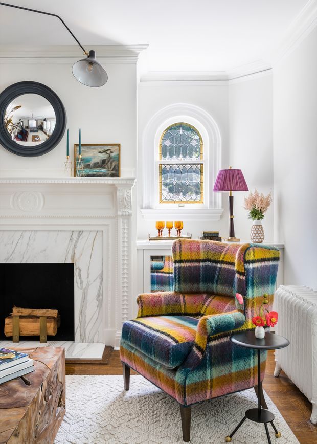Bright, refined living room with white ornate architectural detailing, a marble fireplace surround, arched stained-glass window, dark circular mirror, and a vividly multicoloured plaid armchair. A sculptural floor lamp, side table, radiator, artwork, and small floral arrangement complete the eclectic yet elegant interior.