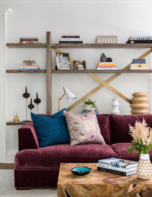 A richly styled living room with a deep burgundy velvet sectional sofa, jewel-toned blue and patterned cushions, and a rustic timber coffee table. Behind the sofa, open wooden shelving displays books and decorative objects, with diagonal timber bracing creating a distinctive feature wall. A floor lamp, ceramics, plants, and floral arrangements add layered detail to the warm, eclectic space.
