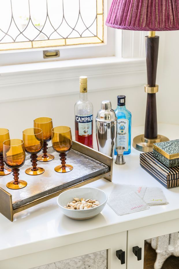 A styled drinks station on a white built-in cabinet beneath a decorative leaded-glass window. The scene includes a silver serving tray with amber cocktail glasses, liquor bottles, a cocktail shaker, a dark wood and brass table lamp with a magenta shade, stacked books, and a small bowl. The cabinetry has dark handles and textured light-coloured drawer fronts, creating a refined vintage-inspired interior vignette.