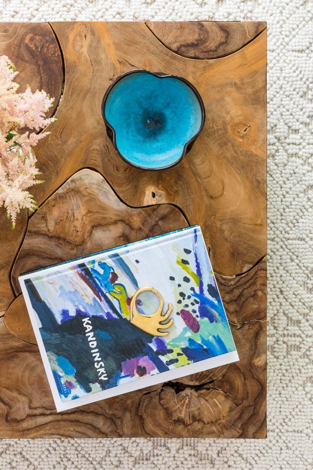 Top-down styled still life showing a rustic live-edge timber coffee table with prominent natural grain and sculpted organic contours. A bright turquoise ceramic bowl, a colourful art book, and pale dried flowers sit on the table, which is surrounded by a textured off-white rug.
