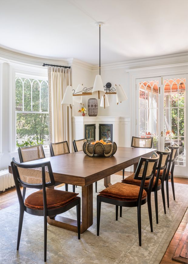 Bright, elegant dining room with a large dark timber rectangular dining table surrounded by black-framed cane-back chairs with brown leather seats. The room features white painted walls, ornate trim and cornices, a decorative fireplace with built-in cabinetry, tall arched windows and glazed French doors, a sculptural multi-light pendant, and a large neutral rug over timber flooring.