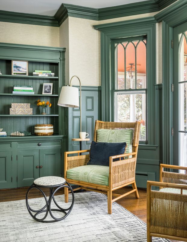 A richly detailed traditional sitting room with deep green built-in bookcases and cabinetry, paneled walls, ornate trim, and tall leaded-glass windows. A woven rattan armchair with green patterned cushions sits beside a small round black metal table, with additional woven seating in the foreground. Warm timber flooring, a pale textured rug, and layered decorative objects create a refined, inviting atmosphere.
