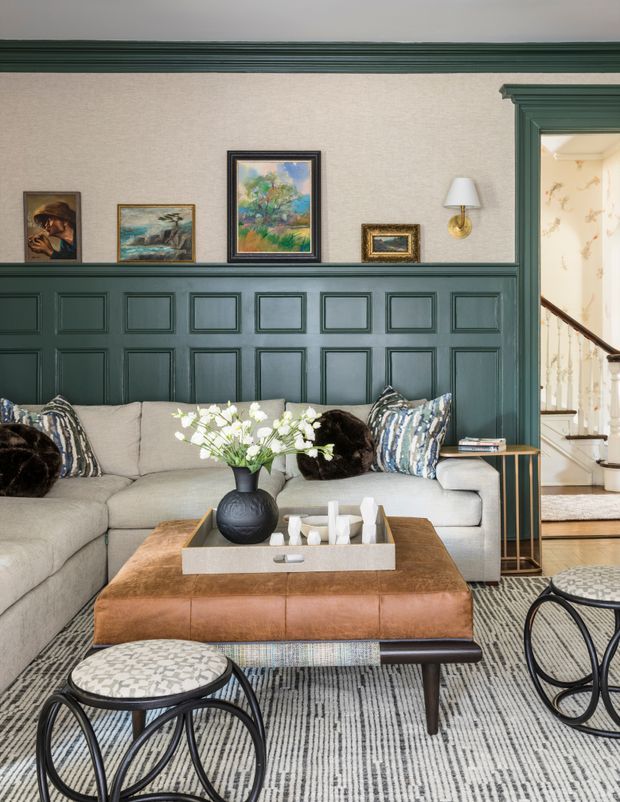 A refined living room with a large light sectional sofa, leather ottoman coffee table, patterned rug, decorative stools, framed artwork, and a dark green painted panelled wall with traditional moulding. A staircase and adjoining space are visible through the doorway.