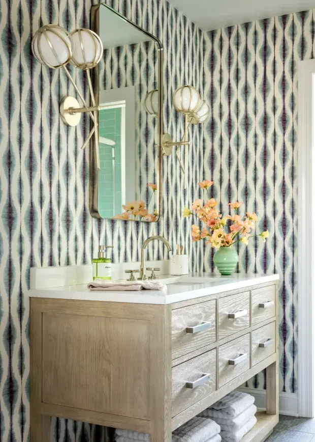 A bright, highly styled bathroom vanity area featuring a light timber floating-style console with a white countertop, integrated drawers, open towel storage, and a curved chrome faucet. The walls are covered in bold teal, white, and dark patterned wallpaper, complemented by a large rounded-rectangle mirror and sculptural globe wall sconces in brass and white. Floral arrangements and neatly folded towels add decorative detail.