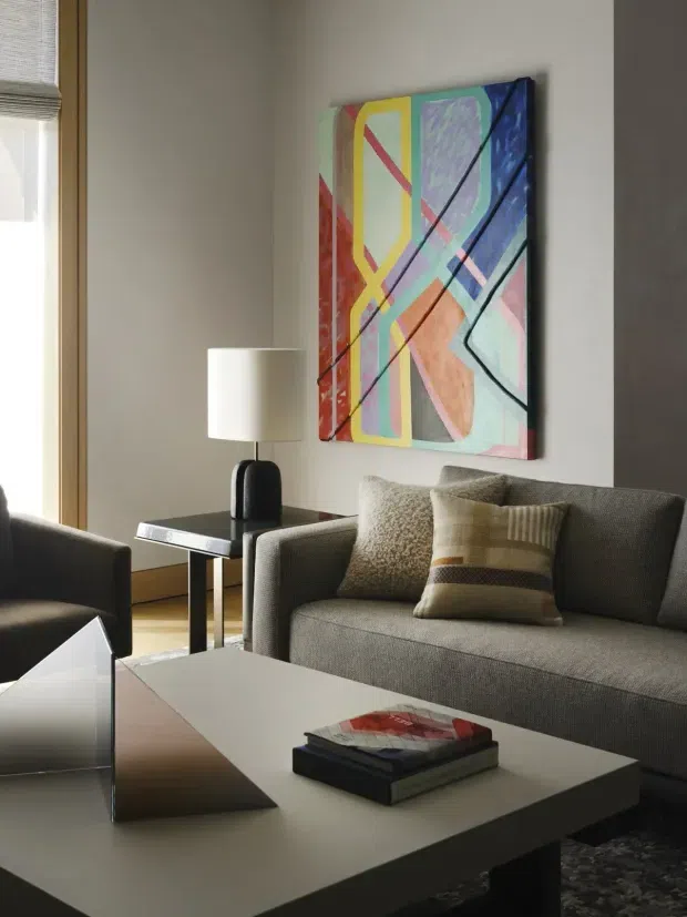 A refined living room with a grey upholstered sofa and armchair arranged around a large rectangular coffee table. A colourful abstract artwork hangs above the sofa, while a sculptural table lamp, books, cushions, and a patterned rug add detail. Natural light enters through a tall window with a roller blind and warm timber trim. The room has a calm, contemporary feel with neutral furnishings and bold artistic accents.