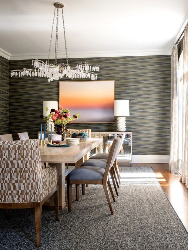 A refined dining room with a long timber dining table, upholstered dining chairs, patterned armchairs, and a large textured rug. The room features dramatic geometric dark wallpaper, white decorative ceiling moulding, full-height sheer curtains, a crystal chandelier, matching table lamps, a large abstract artwork, and a mirrored sideboard. Bright natural light enters from the right, creating a polished, layered interior.