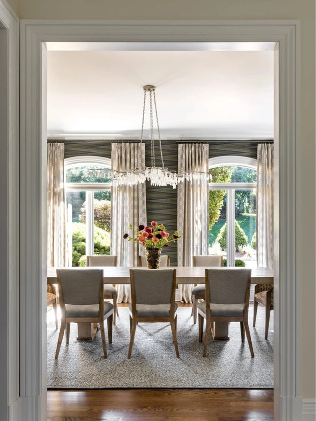 Elegant formal dining room viewed through a wide trimmed doorway, featuring a long dining table with upholstered chairs, a crystal chandelier, floor-to-ceiling patterned curtains, large arched windows, dark patterned wallcovering, and a bright landscaped garden beyond.