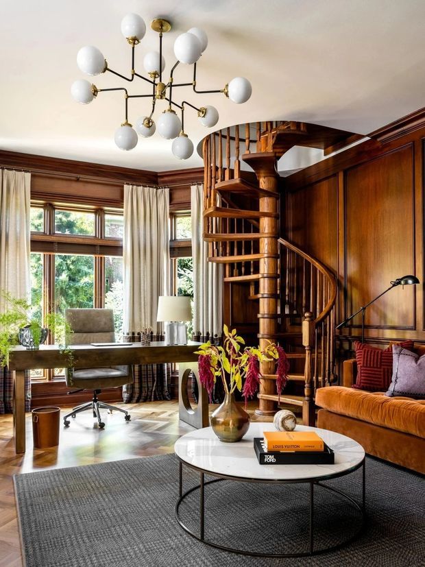 A richly detailed study-lounge interior featuring dark timber wall panelling, a sculptural spiral staircase, tall multi-pane windows with cream curtains, a large timber desk, a leather sofa, and a round marble-topped coffee table. A brass-and-globe chandelier provides a striking focal point, while layered rugs, books, and floral arrangements add warmth and character.