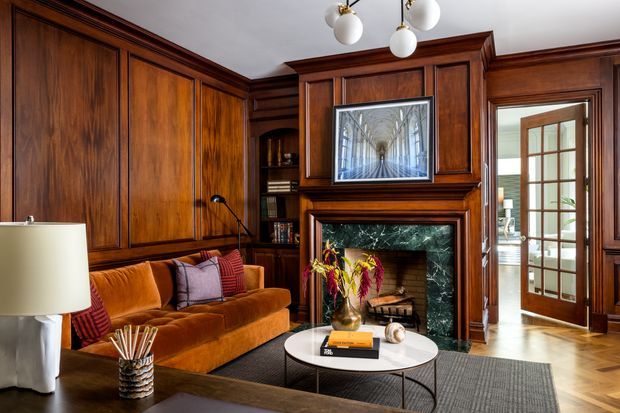 A richly detailed traditional living room with dark walnut-toned timber wall panelling and built-in cabinetry, a marble-surround fireplace, and a warm ochre velvet sofa. A round coffee table, layered rug, decorative lighting, artwork, and a glazed timber door complete the refined, moody interior.