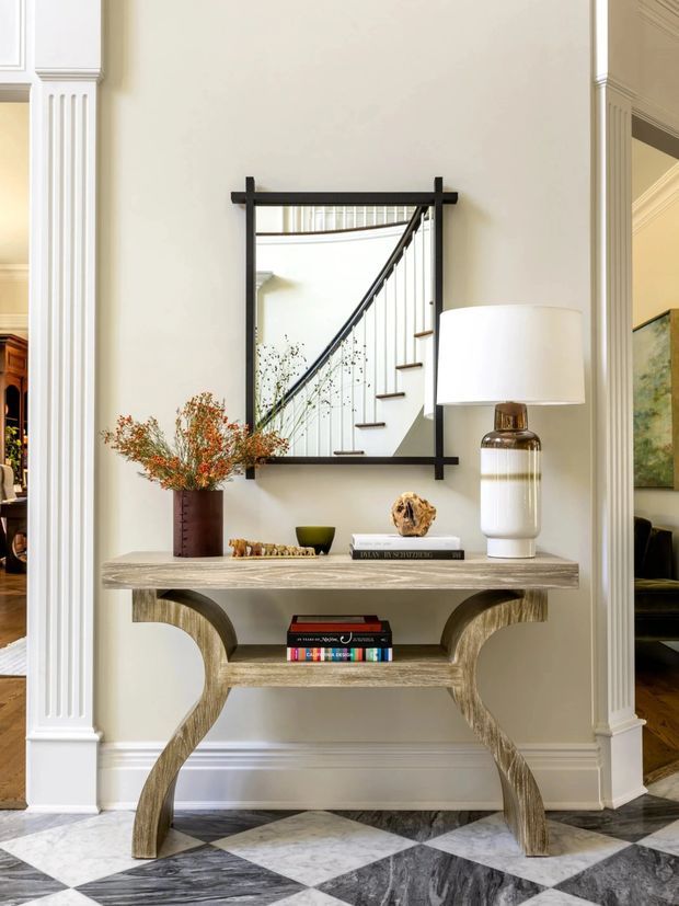 A refined traditional interior vignette featuring a sculptural wood console table against a pale paneled wall, topped with a lamp, vase, books, and decorative objects. A black-framed mirror reflects a staircase with slender balusters. The space has substantial white architectural trim, fluted pilasters, and a dramatic black-and-white geometric stone floor.
