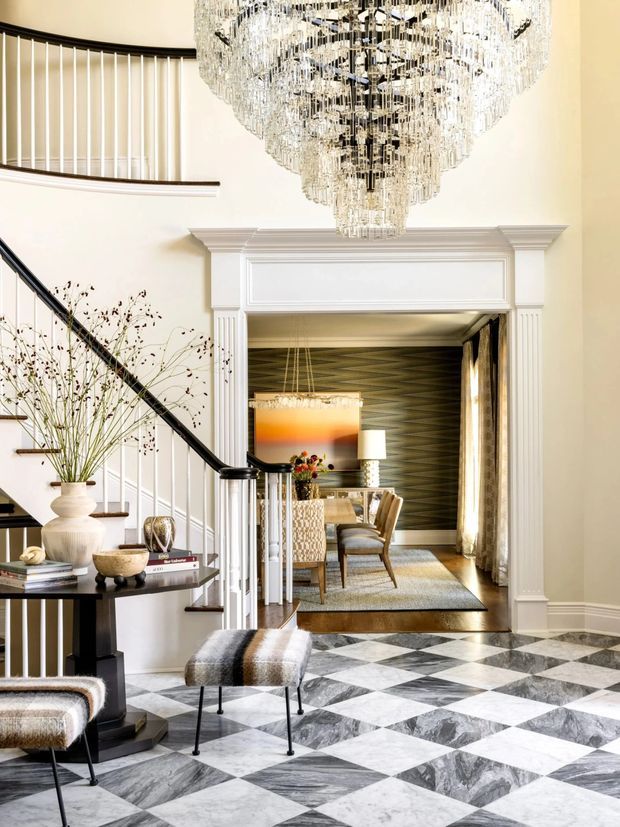 A refined, double-height residential entry hall with a grand crystal chandelier, sweeping staircase, black-and-white marble checkerboard flooring, and white classical wall mouldings. A console table and upholstered stools occupy the foreground, while a formal dining room with patterned dark wallcovering, curtains, and warm lighting is visible through the wide opening.