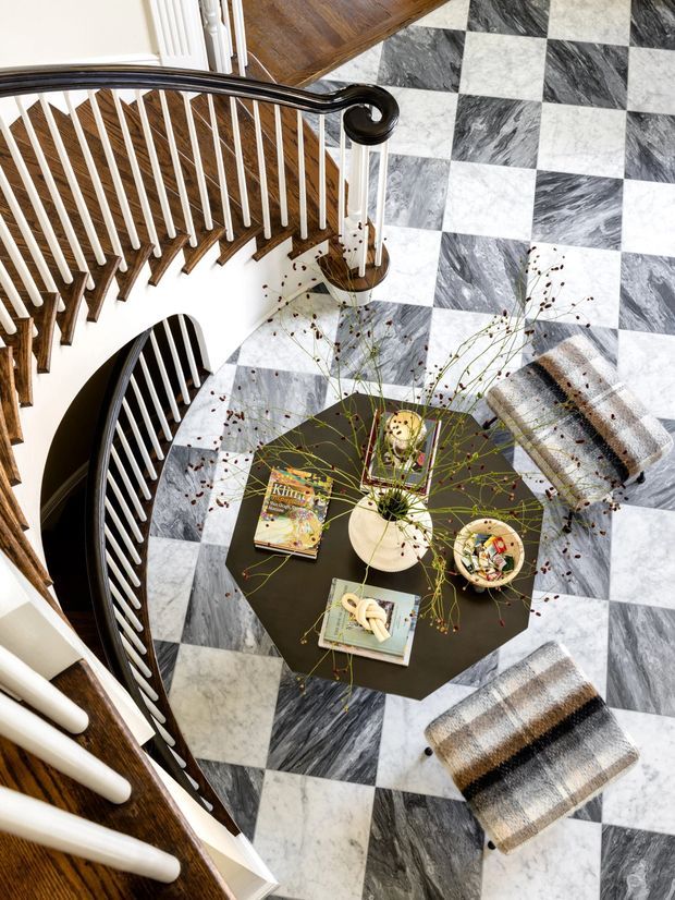 Overhead view of a dramatic curved staircase and landing with dark timber handrails, white balusters, and a bold checkerboard marble floor. A black octagonal coffee table with books, bowls, and dried floral arrangements is surrounded by upholstered stools, creating a refined and graphic transitional interior.