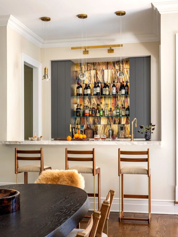 A refined home bar and entertaining area with a marble-topped counter, three timber bar stools, dark built-in cabinetry, open glass shelving, and a striking multicoloured patterned backsplash. Brass pendant and wall lighting, traditional crown moulding, and dark timber flooring create an elegant, warm interior. A round dining table and chairs are visible in the foreground.