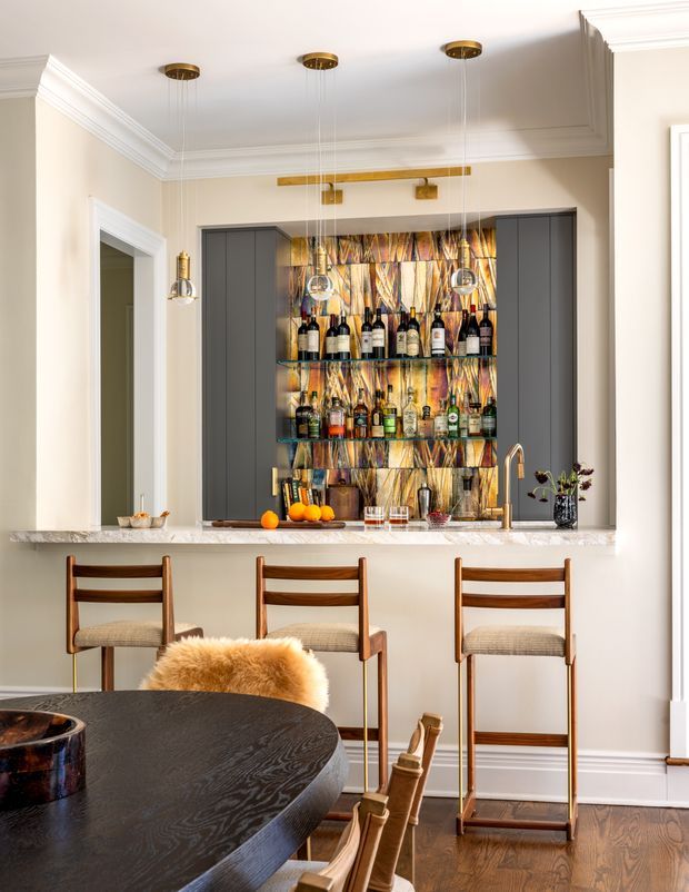 Refined residential home bar and dining area featuring a marble or stone counter, dark grey built-in cabinetry, illuminated glass liquor shelving backed by a colorful patterned feature surface, brass pendant lighting and tapware, and mid-century-inspired wooden bar stools. A dark round dining table and chairs occupy the foreground, with warm timber flooring and classic wall mouldings.