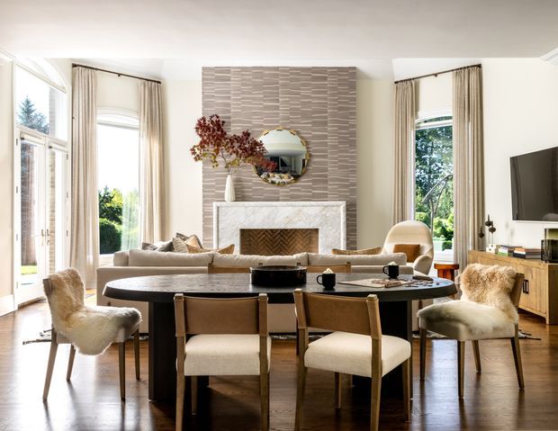 Bright, refined open-plan living and dining room with tall windows, light curtains, a dark oval dining table surrounded by upholstered chairs, and a pale sectional sofa. A marble fireplace with a textured stone feature wall and round mirror forms the focal point, complemented by warm timber flooring and contemporary furnishings.