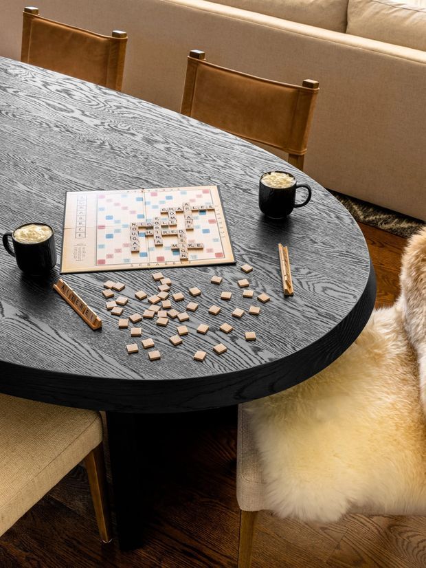A warm, styled dining area centered on a round dark timber dining table with a prominent textured wood-grain surface. The table is set with a Scrabble board, scattered letter tiles, wooden tile racks, and two black mugs of coffee. Upholstered dining chairs, a beige sofa, a wood floor, and a cream sheepskin throw complete the cozy composition.