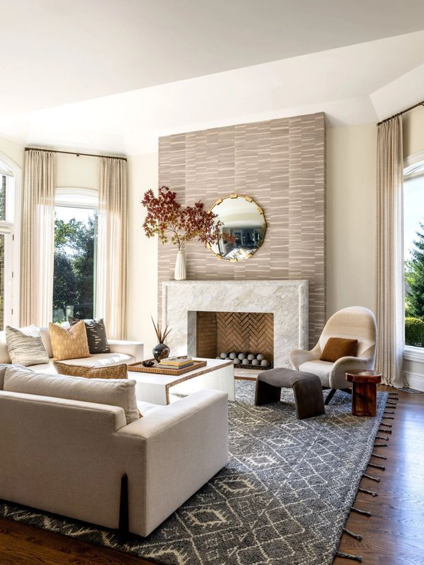 Bright, refined living room with a large upholstered sectional, armchair and ottoman seating arranged around a marble fireplace. The fireplace wall features a textured stacked-stone or tile finish and a round decorative mirror, while tall windows with drapery bring in abundant natural light. Dark timber flooring and a large patterned area rug add contrast.