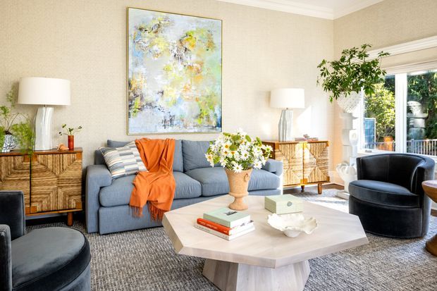 A bright, refined living room with a blue-grey sofa, black accent chairs, a faceted pale wood coffee table, and patterned grey rug. Layered neutral furnishings are complemented by an orange throw, striped cushions, fresh flowers, sculptural lamps, timber sideboards, and a large abstract artwork. Glazed doors open to greenery outside.