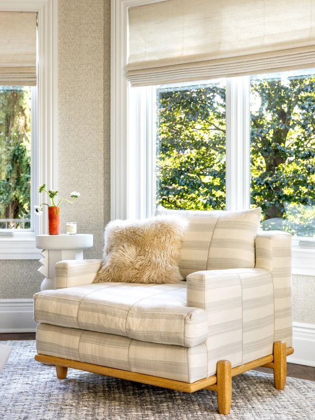 Bright, traditional-style sitting area featuring a large upholstered armchair with matching ottoman, striped cream fabric, a shaggy accent cushion, and exposed light timber legs. The chair sits beside a small white pedestal side table with a vase, in front of tall bay-style windows with woven Roman blinds. Textured neutral wallpaper, white trim, and a patterned rug complete the calm, light-filled setting.
