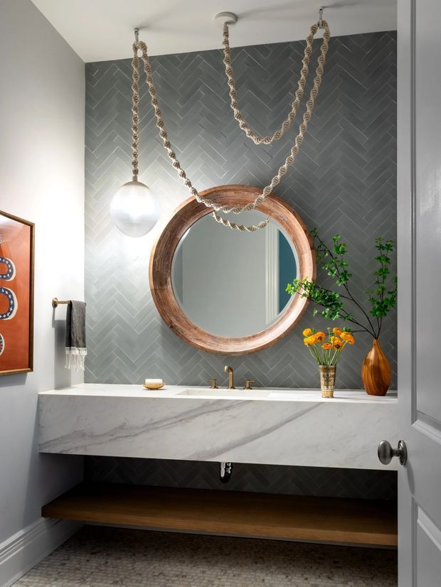 Stylish bathroom vanity with a floating white marble benchtop, dark grey herringbone tiled feature wall, round timber-framed mirror, brass tapware, rope-hung pendant lighting, and an open dark timber shelf beneath. Decorative plants and warm accessories add contrast to the refined monochromatic scheme.