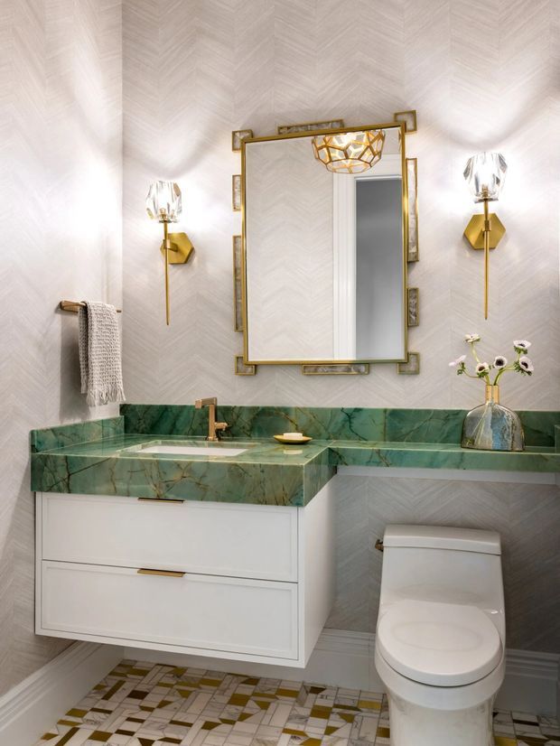 A refined bathroom featuring a floating white vanity with brass hardware, a rich green veined stone benchtop and backsplash, patterned geometric floor tiles, pale herringbone wall finish, brass sconces, and an ornate gold-framed mirror. A toilet and decorative vessel are also visible.