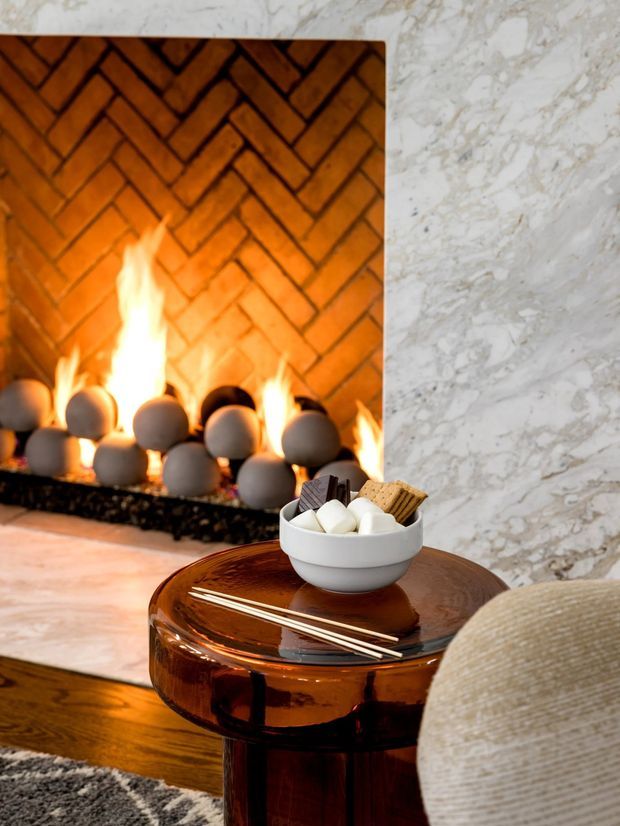 Close-up interior vignette of a warm contemporary living room featuring a lit fireplace lined with a herringbone-patterned terracotta brick surround and framed by veined white marble. A glossy dark amber-brown round side table holds a bowl of marshmallows and crackers, with a textured upholstered chair and dark rug completing the cozy setting.