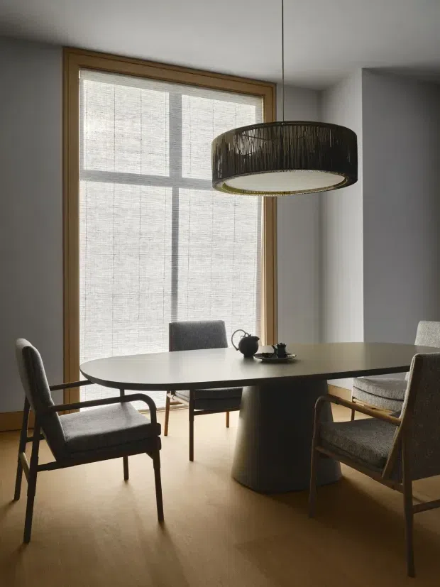 A softly lit dining room with a round dining table surrounded by upholstered armchairs, a small tea setting, and a large woven pendant light. The room features warm timber flooring, a timber-framed window with a translucent roller blind, and restrained neutral furnishings in a calm, refined setting.