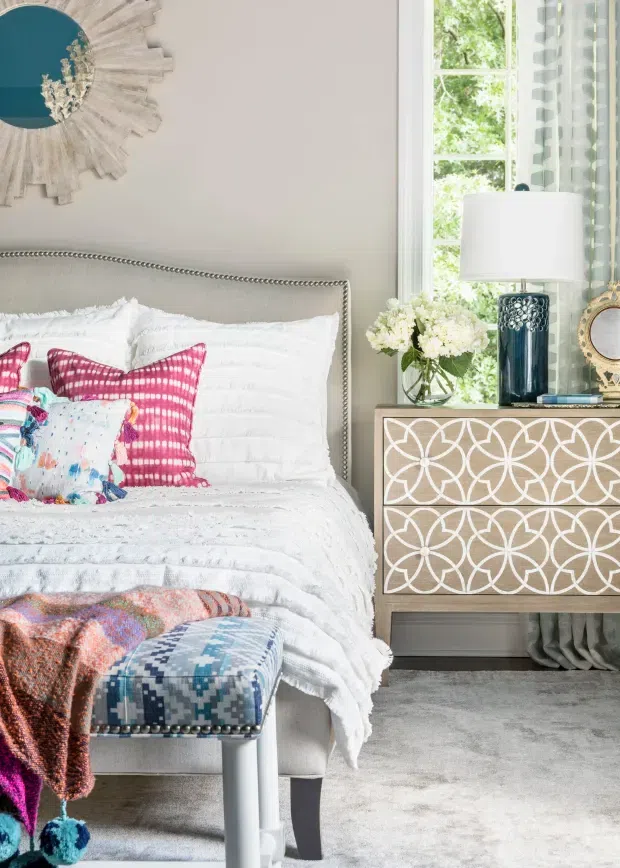 Bright, richly layered bedroom vignette featuring a light upholstered bed with nailhead trim, white textured bedding, vivid pink and multicoloured accent cushions, and a patterned upholstered bench. A decorative geometric-front wooden dresser sits beside the bed beneath a window, styled with a blue ceramic lamp, flowers, and a round mirror. The room has soft neutral walls, grey carpet, patterned curtains, and eclectic colourful detailing.