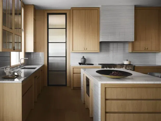 A warm, refined kitchen with extensive light timber cabinetry, a large island, pale tiled surfaces, and a black-framed glazed door. The compact galley-style layout includes a sink beneath a window, integrated storage, a cooktop, and a tiled rangehood surround. The island has a pale stone or composite benchtop and timber drawer fronts, while the dark timber floor adds contrast.