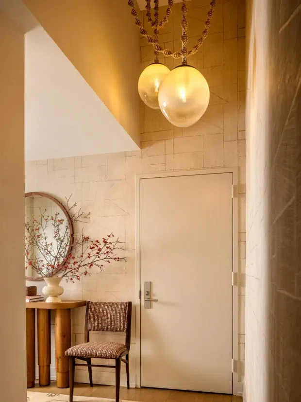 Warmly lit interior entry area with a closed flush door, textured light stone or tile wall finish, sculptural pendant lights, a round wall mirror, narrow timber console table, decorative vase and branch arrangement, and a woven accent chair. The space has a refined, softly layered contemporary aesthetic with warm neutral tones.