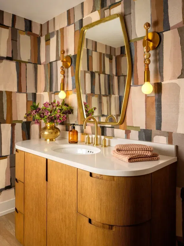 A warmly lit bathroom vanity featuring a curved white countertop with integrated oval basin, rich mid-tone timber cabinetry, brass-gold tapware, and a large organically shaped brass-framed mirror. Sculptural brass wall sconces with exposed globe bulbs flank the mirror, while bold geometric multicoloured wallpaper creates a distinctive feature wall. Decorative flowers, amber toiletries, and folded towels add styling.