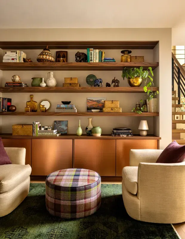Warmly styled living room with a built-in timber shelving and cabinetry wall, displaying books, pottery, art objects and plants. Two cream upholstered armchairs face a rounded plaid ottoman over a dark patterned rug. A timber stairway with black metal balustrading is visible at the right edge.