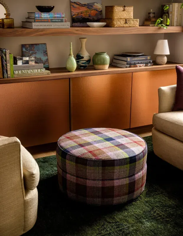 A warm, richly styled living room vignette featuring a built-in timber shelving and cabinetry unit, decorated with books, pottery, woven baskets, artwork and a table lamp. A large plaid upholstered ottoman sits centrally on a deep green rug, with cream upholstered armchairs partially visible at either side. The space has an intimate, layered and refined mid-century-inspired character.